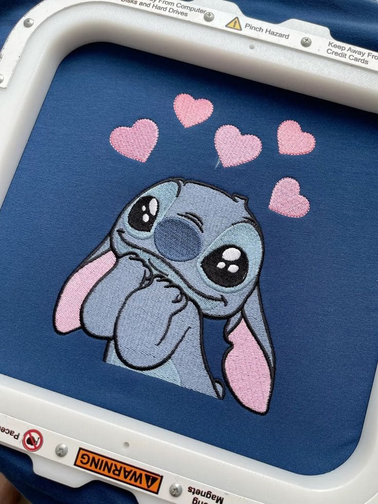 Gestickte Stitch-Figur mit fünf rosa Herzen auf marineblauem Stoff, im Stickrahmen